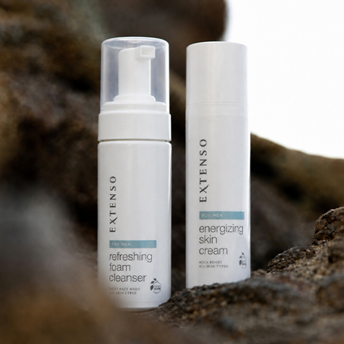 Twee witte Extenso For Men flacons, een schuimende reiniger en een energizing gezichtscrème, gepresenteerd op donkere, ruwe stenen in natuurlijk daglicht.