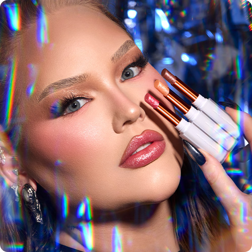 Close-up van NikkieTutorials met glanzende lippenstift, die drie Nimya Discosphere Glitter Lip Toppers vasthoudt tegen een achtergrond met een regenboog-glimmsfeer.