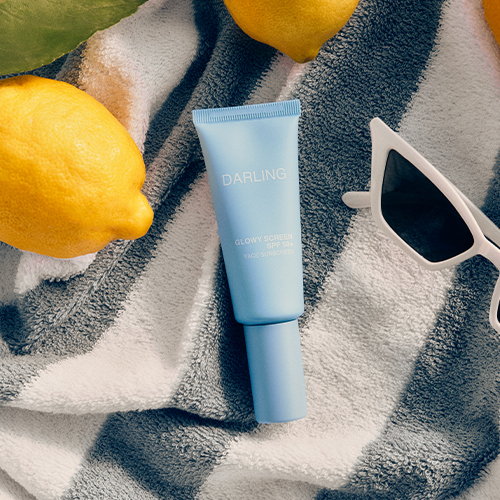 De lichtblauwe tube van Darling Glowy Screen op een gestreepte handdoek naast citroenen en een zonnebril.