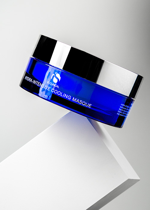 Een blauw glazen potje iS Clinical Hydra-Intensive Cooling Masque dat in balans staat op de hoek van een wit blok tegen een grijze achtergrond.