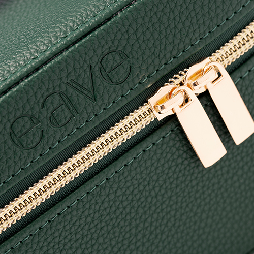 Close-up van de textuur van de groene EAVE vanity bag, met focus op het subtiele reliëf-logo en de hoogwaardige gouden ritssluiting.