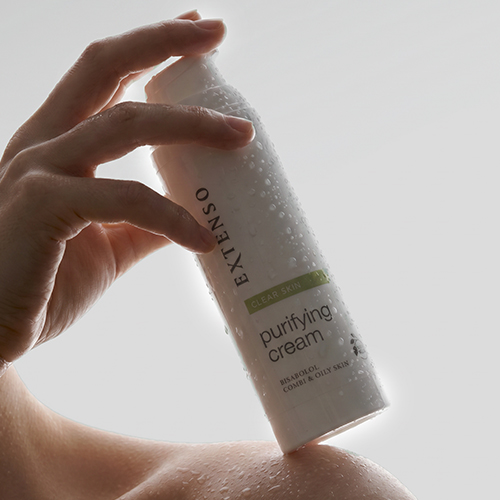 Hand die een flacon Extenso Purifying Cream vasthoudt, bedekt met waterdruppels.