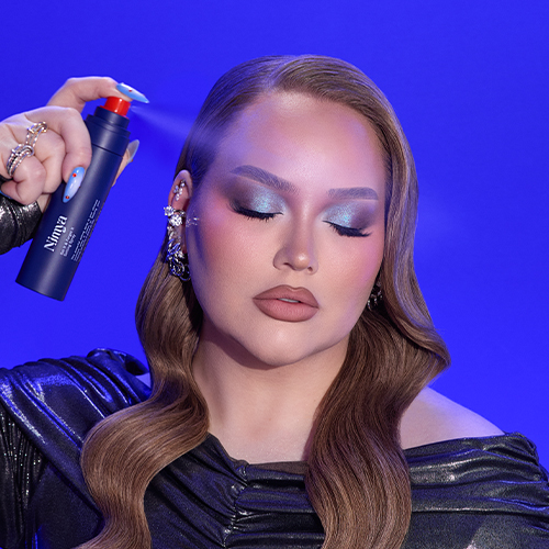 NikkieTutorials sproeit Nimya setting spray op haar gezicht met de blauwe flacon en rode sproeikop tegen een blauwe achtergrond.
