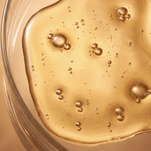 Close-up van de gouden, vloeibare textuur met kleine luchtbelletjes van de gezichtsolie in een glazen schaaltje.