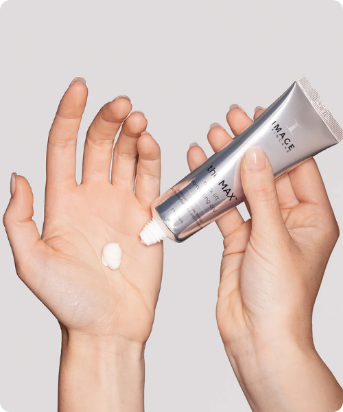 Handen die een kleine hoeveelheid witte IMAGE Skincare the MAX stem cell neck lift crème uit een zilveren tube op de handpalm aanbrengen.