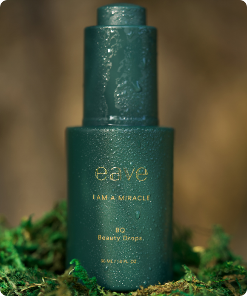 Een donkergroene flacon van EAVE 'I AM A MIRACLE' Beauty Drops, bedekt met waterdruppels en geplaatst op een bed van groen mos tegen een natuurlijke achtergrond.