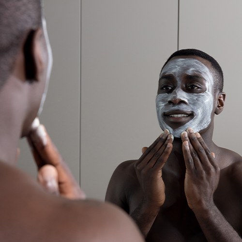 Een man kijkt in de spiegel terwijl hij het witte Synergie Skin Detox anTi ox gezichtsmasker met beide handen zorgvuldig aanbrengt op zijn gezicht.