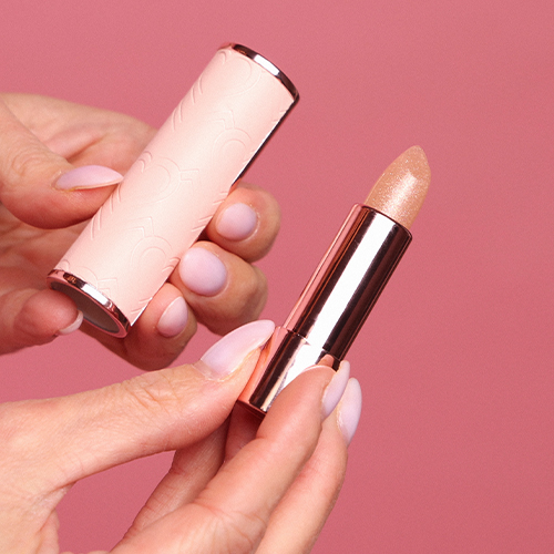 Een foto van de handen van een vrouw met roze nagellak, die een gesloten Cent Pur Cent-lippenstift in rose goud vasthoudt tegen een roze achtergrond.