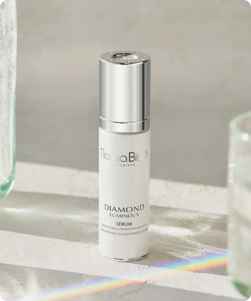 Een flesje Natura Bissé Diamond Luminous Brightening Concentrated Serum op een witte ondergrond, met een subtiele regenboogreflectie van licht op de voorgrond.