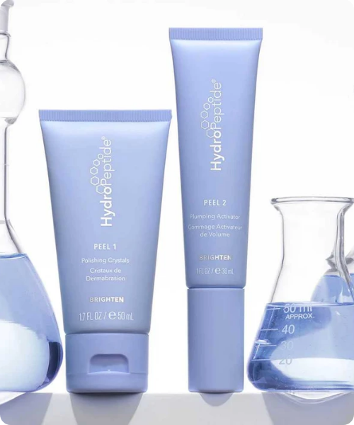 Een productfoto op een witte achtergrond toont twee lavendelkleurige tubes van het merk HydroPeptide, geflankeerd door twee glazen laboratoriumkolven gevuld met een blauwe vloeistof.