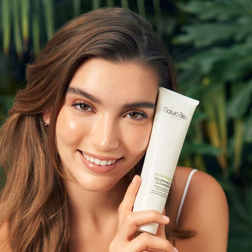Een glimlachende vrouw houdt de Natura Bissé Tolerance Cleanser tube tegen haar gezicht met een achtergrond van groene bladeren.