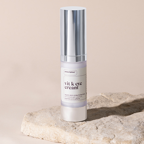 Een flesje Prescription Vit K Eye Cream met zilveren dop rechtopstaand op een ruwe lichte steen tegen een beige achtergrond