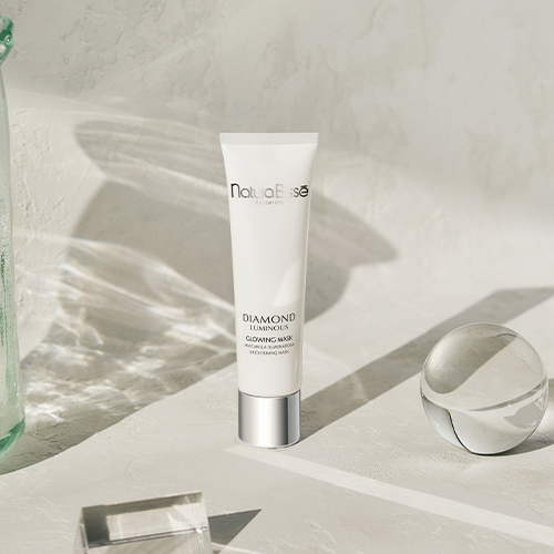 Een staande tube Natura Bissé Diamond Luminous Glowing Mask op een verhoogd platform van getextureerd cement, vergezeld door een glazen karaf en twee glazen bollen onder felle zonneschijn.