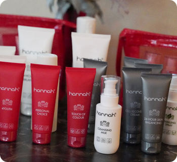 Een overzichtsfoto mobiel van diverse hannah-huidverzorgingsproducten, zoals crèmes, serums en lotions, gepresenteerd in rode en witte verpakkingen op een donkere tafel.