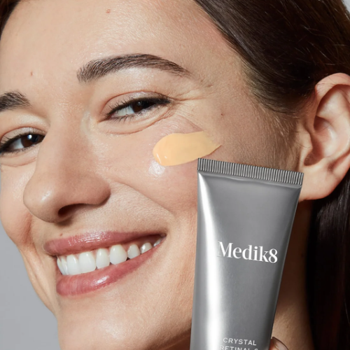 Een glimlachende vrouw met een veeg gele crème op haar wang, die een zilveren tube Medik8 Crystal Retinal vasthoudt.