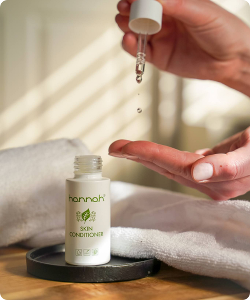 Een hand met donker gelakte nagels houdt een wit flesje hannah Skin Conditioner vast tegen een witte, gestructureerde achtergrond.