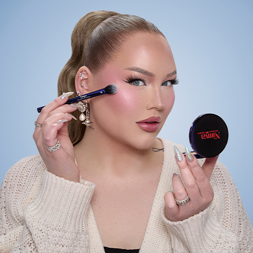 NikkieTutorials brengt met een blauwe Nimya kwast highlighter aan op haar jukbeen en houdt een zwart spiegeltje vast.