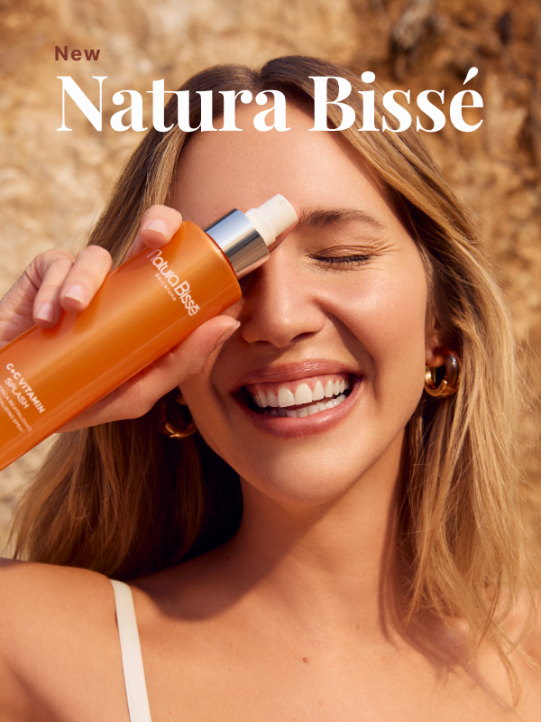 Een lachende vrouw houdt een oranje flacon van Natura Bissé C+C Vitamin Splash voor haar oog tegen een zonnige, rotsachtige achtergrond. Bovenaan staat de tekst "Nieuw Natura Bissé"