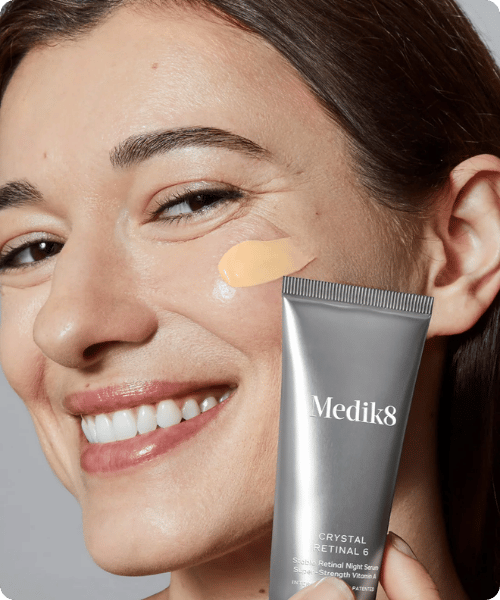 Een glimlachende vrouw brengt een geelkleurig retinol serum aan op haar gezicht met een zilveren tube