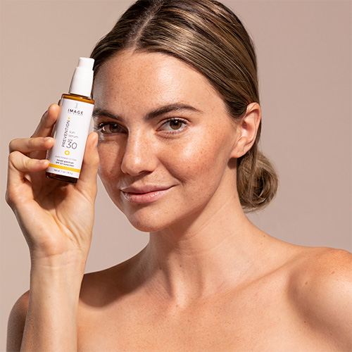 Een vrouw met een stralende huid houdt het IMAGE Skincare Sun Serum SPF 30 vast naast haar gezicht.