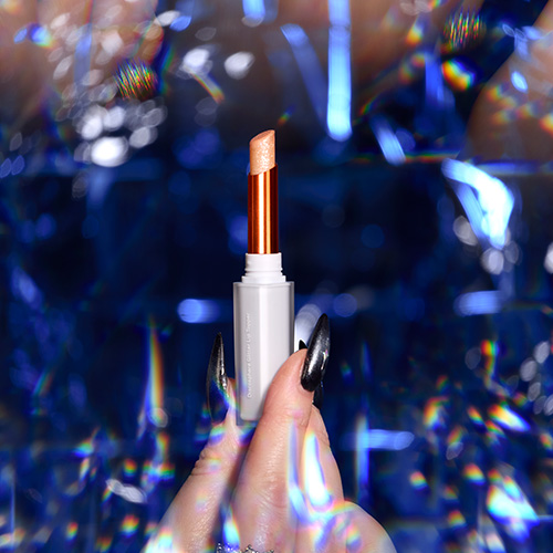 Hand met donkere nagels houdt de open Nimya glitter lippenstift in de kleur Dressed In Diamonds vast voor een blauwe holografische achtergrond