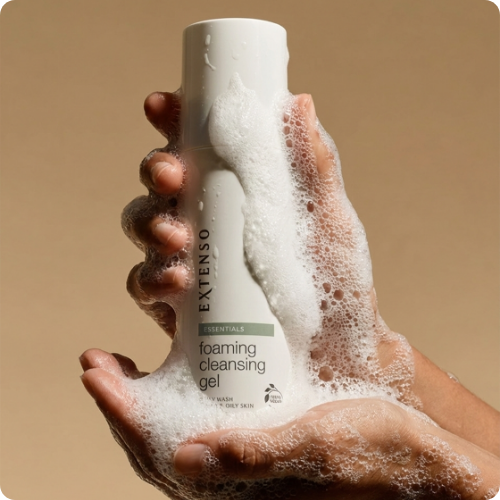 Twee handen vol met rijk wit schuim houden de Extenso Essentials foaming cleansing gel flacon vast tegen een egale beige achtergrond.