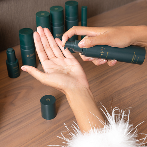 Een persoon pompt de Eave I AM PURE Face Cleanser in hun handpalm. In de achtergrond staan andere Eave-producten en een afgehaalde dop. De persoon draagt een witte top met veren.