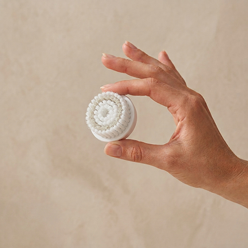 Hand houdt een Extenso exfoliërende opzetborstel vast tegen een beige achtergrond