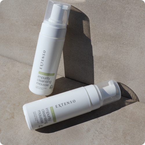 Twee flacons van de Extenso Clear Skin impurity cleansing mousse liggen op een zandkleurige, stenen ondergrond in de schaduw en het zonlicht.