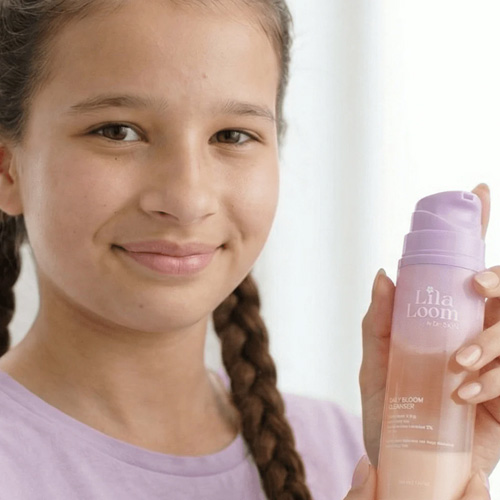 Een meisje met vlechten houdt de Lila Loom Daily Bloom Cleanser vast, een doorzichtige flacon met paarse details.