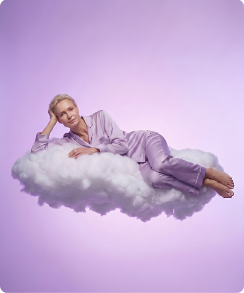Een vrouw met kort blond haar draagt een paarse satijnen pyjama en ligt ontspannen op een witte wolk tegen een paarse achtergrond