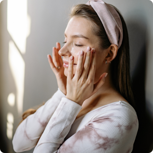 Vrouw met een roze haarband brengt zachtjes een verzorgingsproduct aan op haar gezicht met gesloten ogen in zacht daglicht.