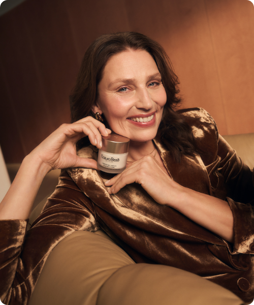 Een lachende vrouw met golvend bruin haar zit op een leren bank en houdt een potje Natura Bissé Essential Shock Intense Cream dicht bij haar gezicht. Ze draagt een bruine, glanzende fluwelen blazer tegen een warme, houten panelen achtergrond.