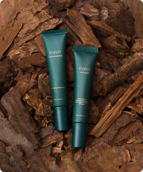 Twee donkergroene tubes van de Eave 'I AM RADIANT' dagcrème en 'I AM REJUVENATING' nachtcrème, gepresenteerd op een ondergrond van natuurlijke bruine boomschors.