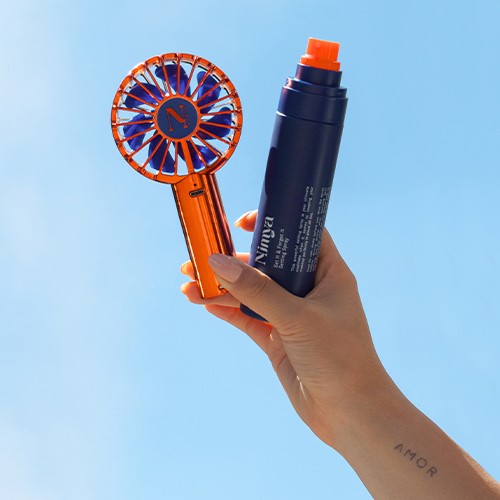 Hand houdt de oranje Nimya ventilator en een blauwe Nimya spray vast tegen een blauwe lucht