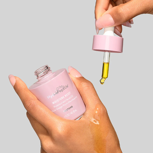 Hand die een roze flesje HydroPeptide Moisture Reset gezichtsolie vasthoudt met een pipet erboven waar een druppel gele olie uit druipt.