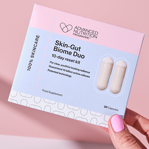 Een hand met roze nagellak houdt een verpakking vast van de Skin Gut Biome Duo resetkit op een roze achtergrond.