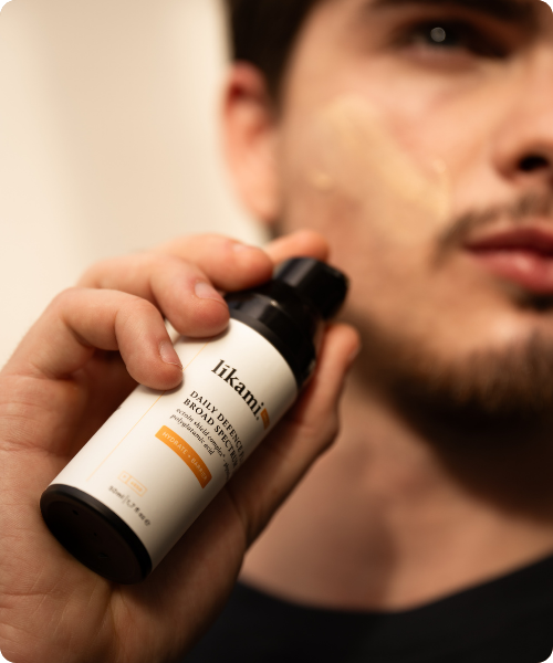 Een persoon houdt een flesje Likami Daily Defence Fluid 'Hydrate + Barrier' vast, terwijl er een kleine hoeveelheid van het product op hun wang is aangebracht.