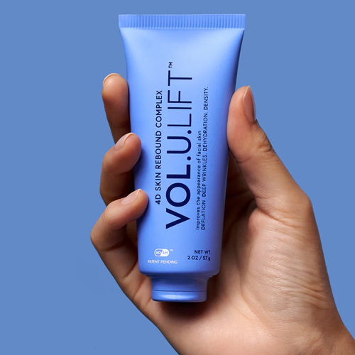 Een hand houdt de blauwe tube van IMAGE Skincare VOL.U.LIFT 4D Skin Rebound Complex vast tegen een blauwe achtergrond.