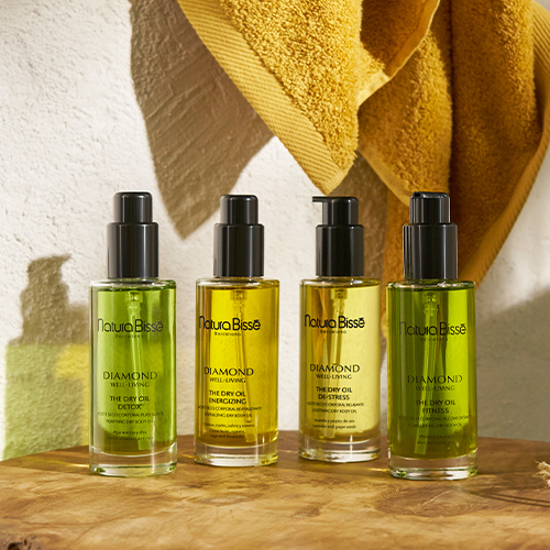 Vier verschillende Natura Bissé Diamond Well Living body oils op een houten ondergrond met een gele handdoek op de achtergrond.
