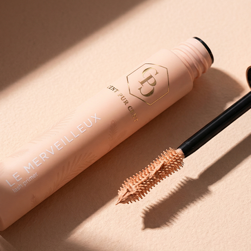 Close-up van de geopende mascara primer op een beige ondergrond, liggend naast het borsteltje dat bedekt is met primer.