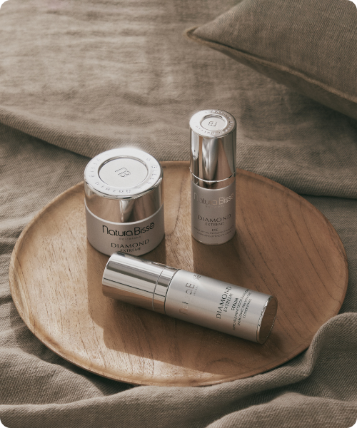 Een zilverkleurige pot en twee flacons van de Natura Bissé Diamond Extreme lijn liggen op een rond houten dienblad dat op een beige linnen textiel rust.