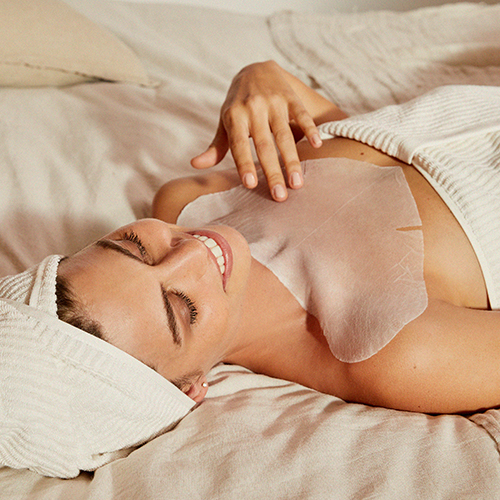 Een liggende vrouw met een wit haarmasker draagt het Tensolift Neck Mask op haar decolleté en hals voor een intensieve behandeling.