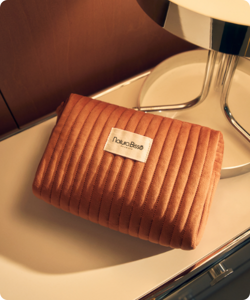 Een gewatteerde oranje bruine fluwelen toilettas met het Natura Bissé label ligt op een witte tafel naast een moderne chroom en witte lamp