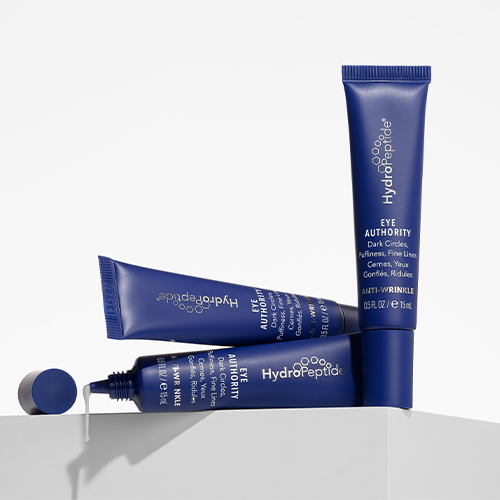 Drie blauwe tubes HydroPeptide Eye Authority oogcrème artistiek opgesteld op een lichte ondergrond waarvan één open is met een druppel crèmepartikel.