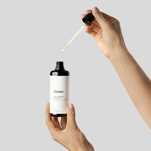 Een close-up tegen een egale grijze achtergrond toont twee handen met een lichte huidskleur en lichtroze nagellak die de Likami Skin Ferment Essence vasthoudt