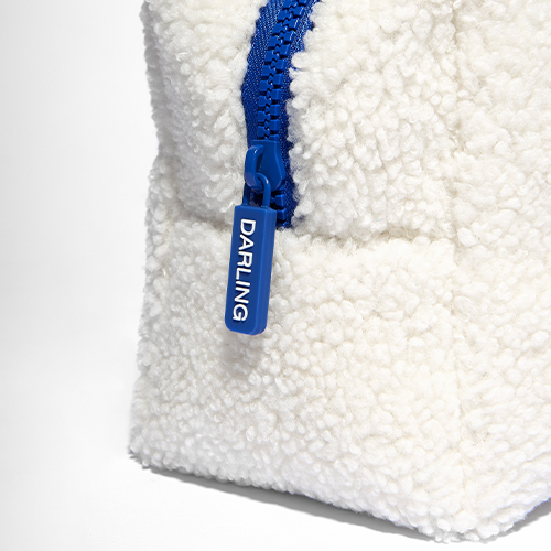 en close-up shot van de witte, pluizige Darling toilettas. De focus ligt op de blauwe rits en het metalen ritslipje waar de merknaam "DARLING" in is gegraveerd.