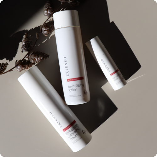 Drie witte flacons van Extenso Skincare uit de anti-aging lijn liggen op een beige ondergrond met harde schaduwen en een gedroogde tak