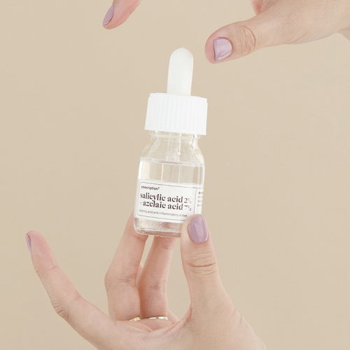 Een close up van twee handen met lila nagellak die een glazen flesje Prescription salicylic acid 2% en azelaic acid 7% vasthouden. De achtergrond is beige.