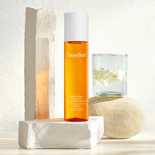 De oranje flacon van het micellair water stijlvol gepresenteerd tussen witte stenen blokken en een glas met witte bloemblaadjes in een heldere, natuurlijke setting.